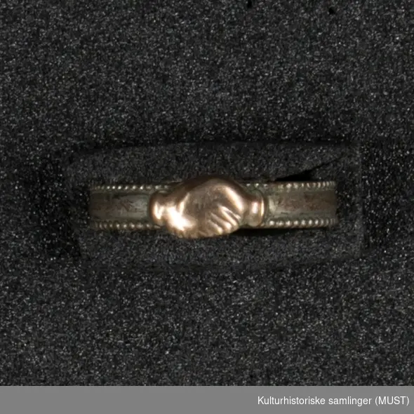 Glatt, flat ring med små, graverte border langs kantene. Påloddet ornament i
kobber bestående av to hender som holder hverandre.