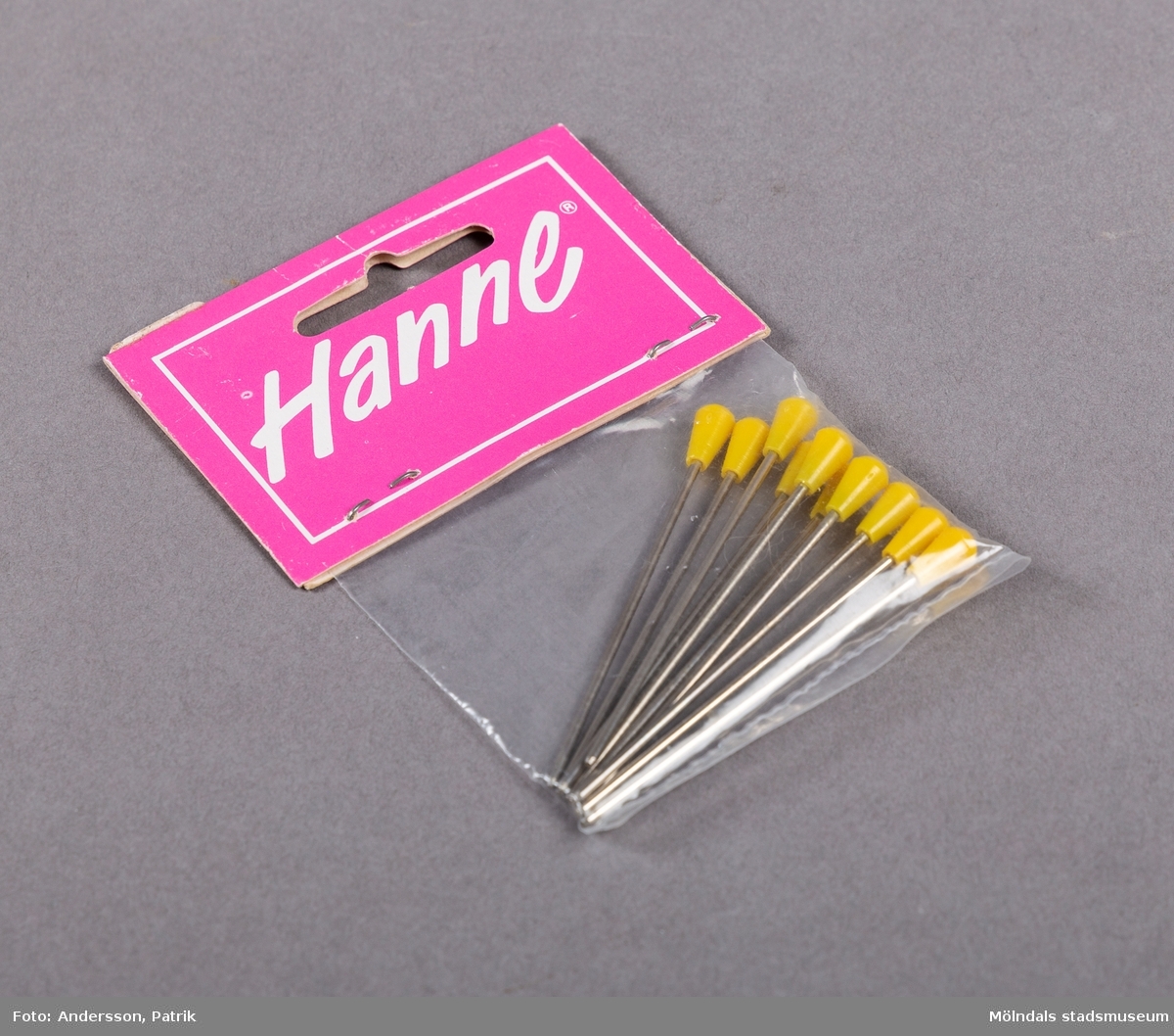 En mindre plastpåse innehållande nålar med gul ände av plast. Etiketten som är rosa, innehåller företagets namn skrivet i vit text, inom en vit ram.
Etiketten är fasthäftad på påsen med två häftstift.