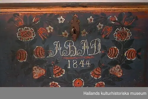 Kista (a) med välvt lock och raka sidor. Kistan är beslagen med i vardera hörn två bandformade hörnbeslag med spetsig avslutning. Nyckelskylt och lås intakt. På locket fem låsbultar, tre stora och två mindre. I sidorna handtag fästade i snedställda kvadrater. Handtagen klena med liten vridning mitt på som dekor. Gångjärn bandformade och raka. Bakre högra har lossnat. Nyckel (b) böjd. Kistan är ommålad. En gammal röd bottenfärg syns genom den senare blå. Locket slitet, endast färgrester. På locket mitt fram en stängel med två rosor med ur blomman uppstående blad och bär. I toppen av stängeln en tulpan. I de främre hörnen på locket fembladig röd blomma med gröna blad med svarta konturer. Lockets list profilhyvlad. På framsidan en symmetrisk krans av blommor. Längst ner i mitten en ros, därefter längs båda stänglarna: rosor, rocailleformad blomma, rosor, rutblomma, tulpaner, rosenknopp och längst upp pingstliljor. Blommor röda med vita detaljer, stängel svart och blad gröna. Rosorna har stängel med blad och rött bär i toppen. Tulpanerna har två långa blad ur blomman. I mitten monogram: "ABAD 1844" i vitt. Texten utformad med samma dekor som broderade monogram. Mellan initialer och årtal ett rött vågigt streck. Baksidan omålad. I främre högra hörnet en liten del av en kistfot bevarad.