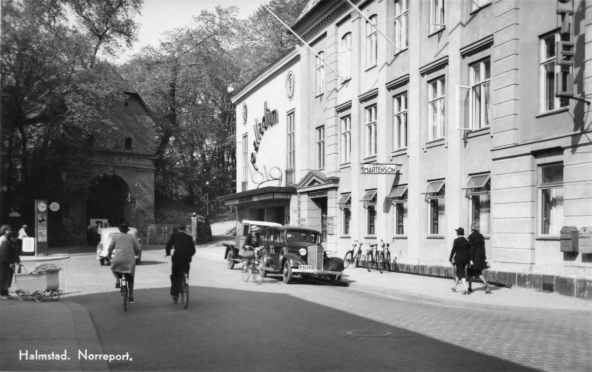 Vykort, "Halmstad. Norreport." Trafikanter och fotgängare på Storgatan vid Norreport. Till höger ligger Bio Palladium och Hotell Mårtenson. Till vänster går ett par damer med barnvagn.