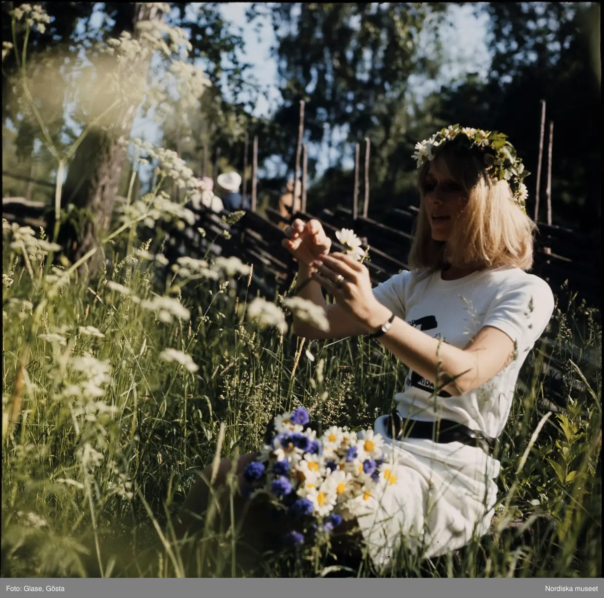 kvinna-sitter-i-gr-set-och-plockar-blommor-med-midsommarkrans-i-h-ret