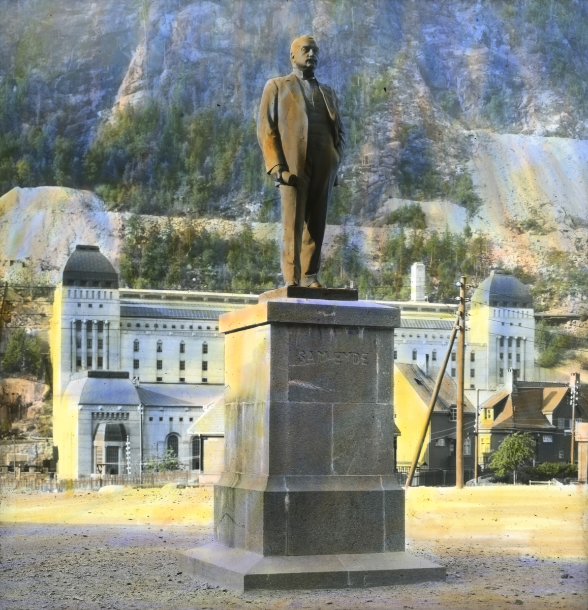 Håndkolorert dias. En statue av Sam Eyde på Rjukan, laget av Gunnar Utsond. Statuen ble avduket på torget i 1920. Bak sees Såheim kraftstasjon.