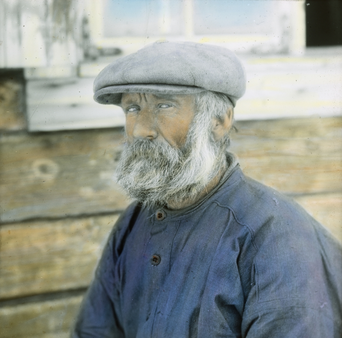 Håndkolorert dias. Portrett av en mann med grått skjegg og hår. Mannen er kledd i blå arbeidsskjorte og grå skyggelue.