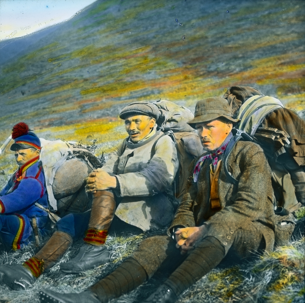 Håndkolorert dias. Tre menn, hvorav en er kledd i samekofte, sitter i en fjellside.