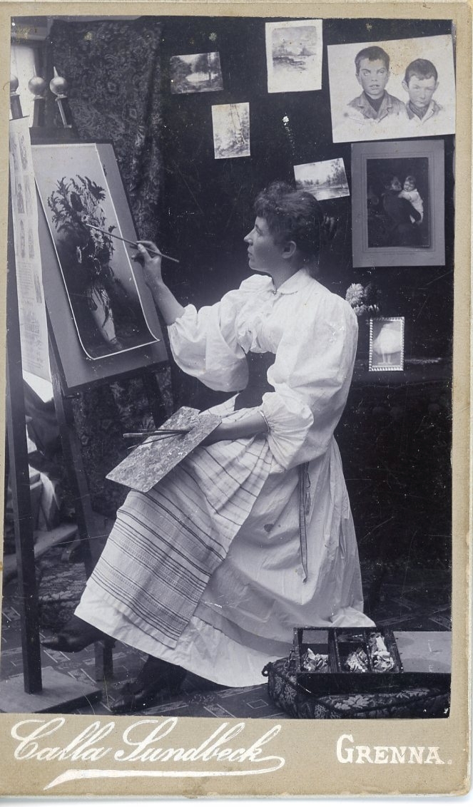 Kabinettsfotografi av en okänd kvinna som sitter vid ett staffli och målar ett blomstermotiv.