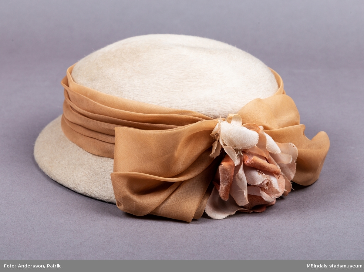 En damhatt i kamelhårsliknande mjukt material. Hatten är ljusbeige i färgen. Den är platt på ovansidan och har ett medelstort brätte. Runt hattens kulle finns ett beigefärgat sidenband som avslutas i en stor rosett på hattens baksida. På rosetten finns en rosa blomma i siden och sammet tyg. 
På insidan har hatten ett ribbsband med en fastsydd etikett. På etiketten finns tillverkarens namn Ateljévåningen Hattbazaren. Den finns ett gummiband fastsytt i hattens ribbsband, för att hålla hatten på plats vid användning.