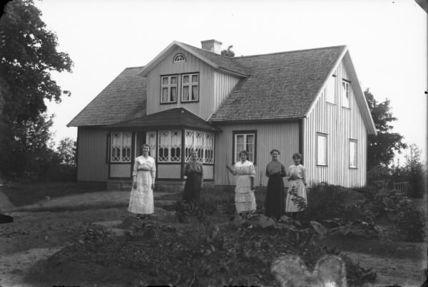 Fem kvinnor står vid trädgårdsodlingen framför ett bostadshus. Huset har en glasveranda och däröver en frontespis upp till nock.