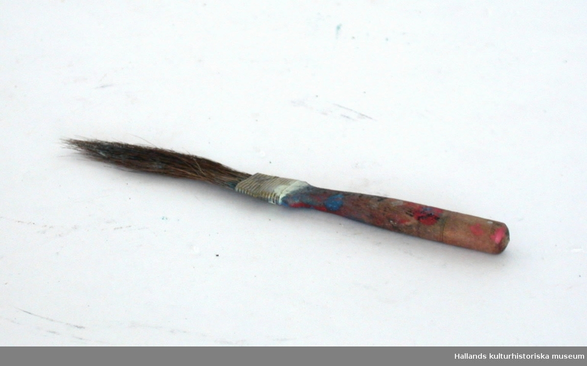 Liten pensel med träskaft. Skaftet smalnar av där "penselhåren", är fastklämda med en metallbit. Blå, röd och vit målarfärg finns på skaftet.