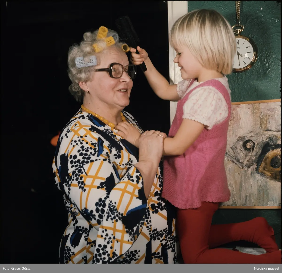 En liten flicka hjälper en äldre kvinna med frisyren. Kvinnan har papiljotter i håret.