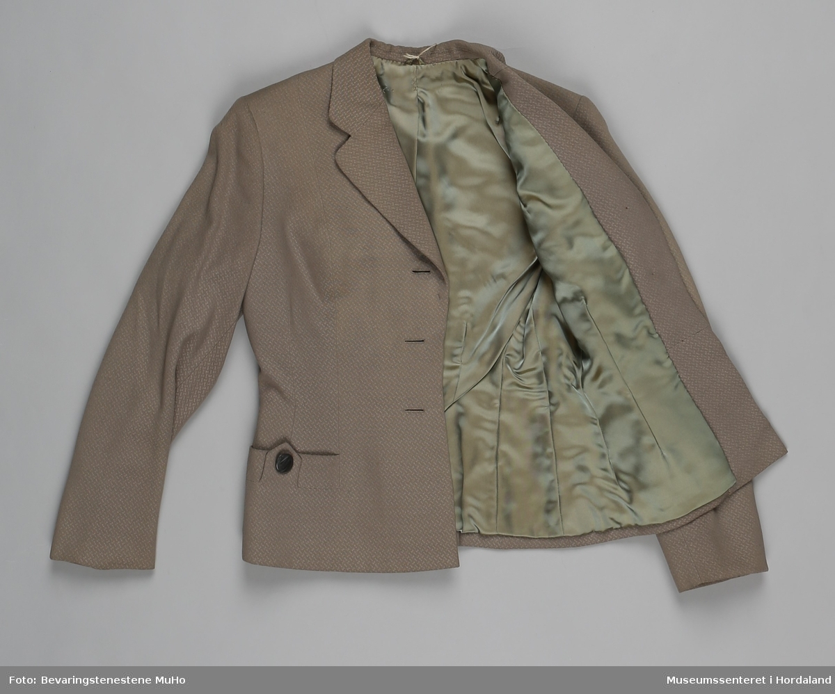 Todelt damedrakt med skjørt og jakke i str. 40. Stoffet er småmønstrete og gråbeige, jakka har brune knappar, knelangt skjørt med strikk og hemper i livet.