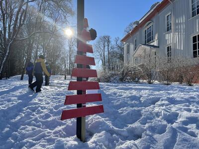 Et vinterbilde viser utsnitt av Eidsvollsbygningens øst-fasade. Sola lyser midt i mot. Noen barn går i snøen i bakgrunnen, i forgrunnen et snekret, rødmalt, grantrelignende installasjon, ca 1,3m høyt. På "treet" henger en natursti-post.