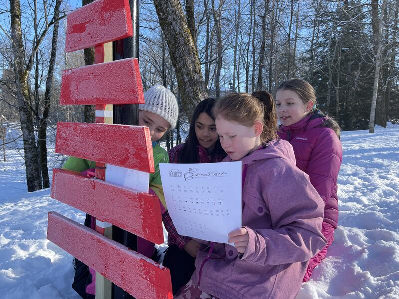 Fire barn i alderen ca 10-12 år sitter på kne i snøen ved en post i vår natursti-løype. De ser konsentrerte ut når de løser oppgaven på arket som henger på posten.
