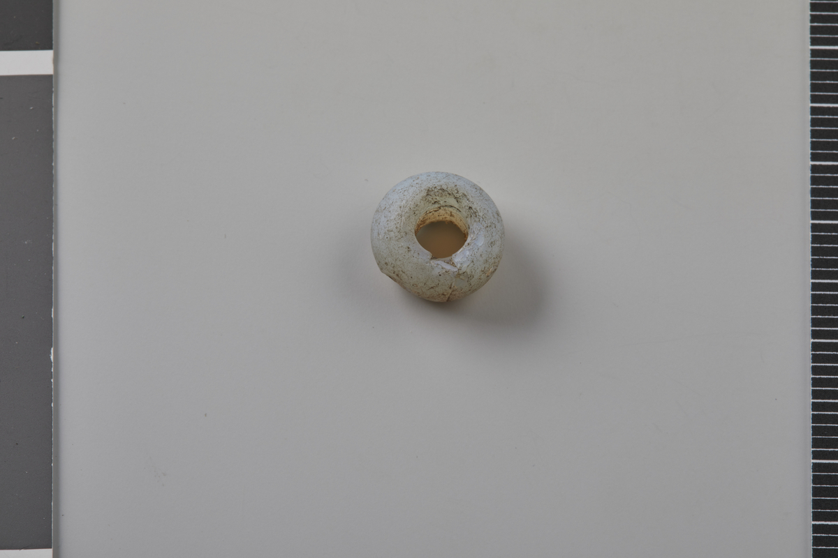 Kuleformet perle av gråhvitt opakt glass, hovedtype som Callmer 1977 A0220. Dateres til yngre jernalder. Diam. 1,1 cm, hullets diam. 0,4 cm, h. 0,7 cm. Vekt: 0,9 g.