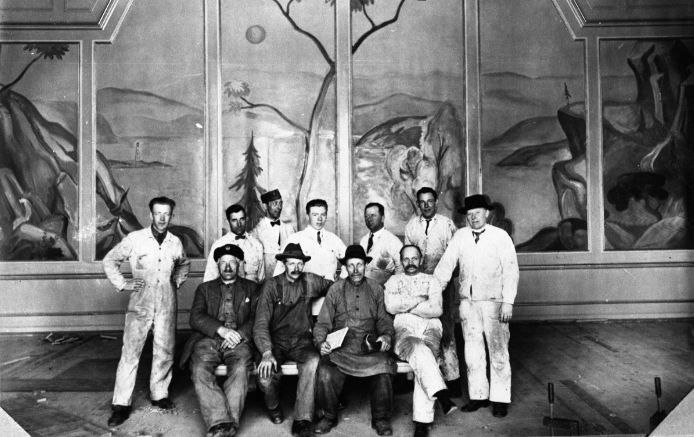 Personalgrupp. En grupp målare och troligen snickare poserar framför en väggmålning indelad i paneler i societetshuset. Sannolikt är det dessa män som skapat landskapsmålningen.