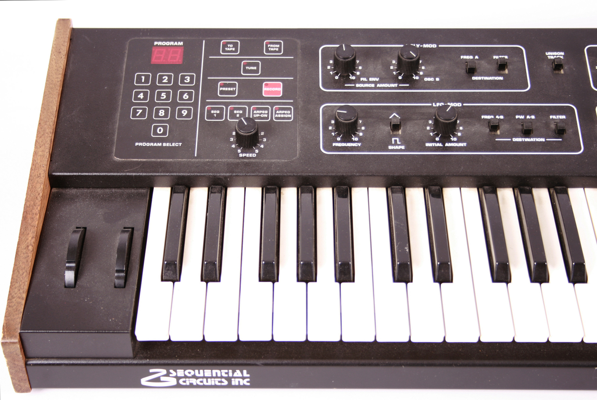 Elektronisk musikkinstrument, synthesizer. Tastatur med klaver nederst mot brukeren. Øverste rekke består av vridbare knapper. På vesntre side et panel med tall. Baksiden har inngang til ulike ledninger.