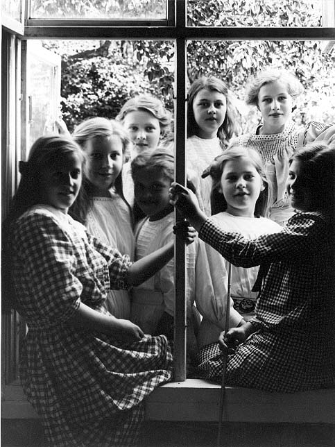 Åtta flickor vid ett fönster till en skolbyggnad, två av dem i rutiga klänningar sitter på fönsterbrädan. Det är sommar.