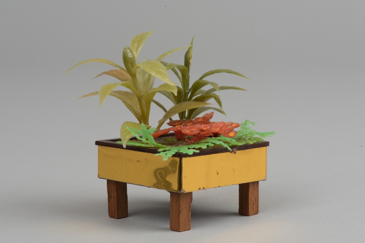 Kvadratiskt blomsterbord med gröna växter, tillverkade i plast.
Bordet har fyra ben av trä, bruna. Sidorna är guldfärgade. 
Inuti bordet står tre olika växter i gröna nyanser och en växt i bruna nyanser.
Bordet är tillverkat av Lundby, som dockskåpstillbehör.