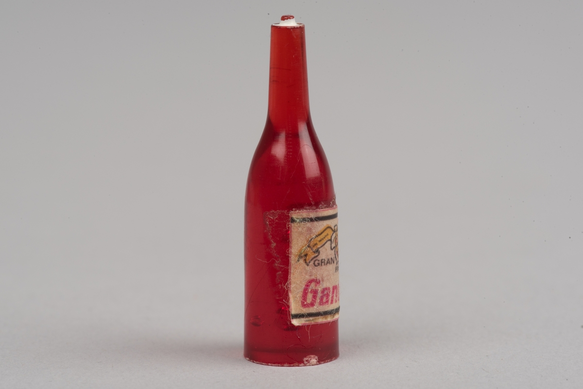 Dockskåpsinredning i form av en vinflaska av plast med etikett.
Flaskan är röd med en vit kork. På etiketten framkommer det att det ska vara en rödvinflaska från Gandesa i Spanien. Etiketten har text och en vindruvsklase.