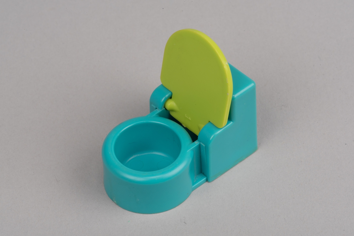 Dockskåpsinredning i form av en toalettstol av plast. Toalettlocket går att fälla upp.
Vattenbehållaren och stolen är turkosa. På ovansidan av behållaren finns en förhöjd spolknapp. Toalettlocket är gulgrönt.