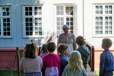 Ei tjenestejente (en ansatt ved museet i kostyme) står ute foran Eidsvollsbygnignen og snakker med en samling barn