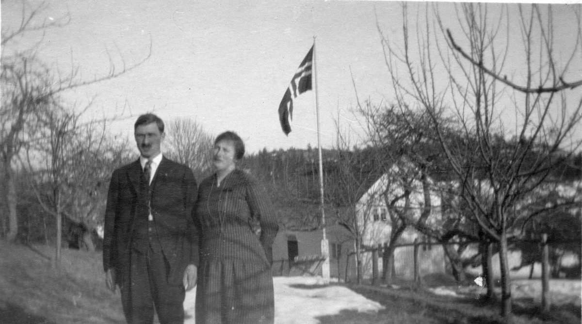 Portrett av mann og kvinne på gårdsvei. Frukthage og det norske flagg i ...