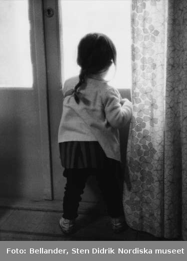 Liten flicka, med ryggen mot fotografen, tittar ut genom ett fönster i en dörr.