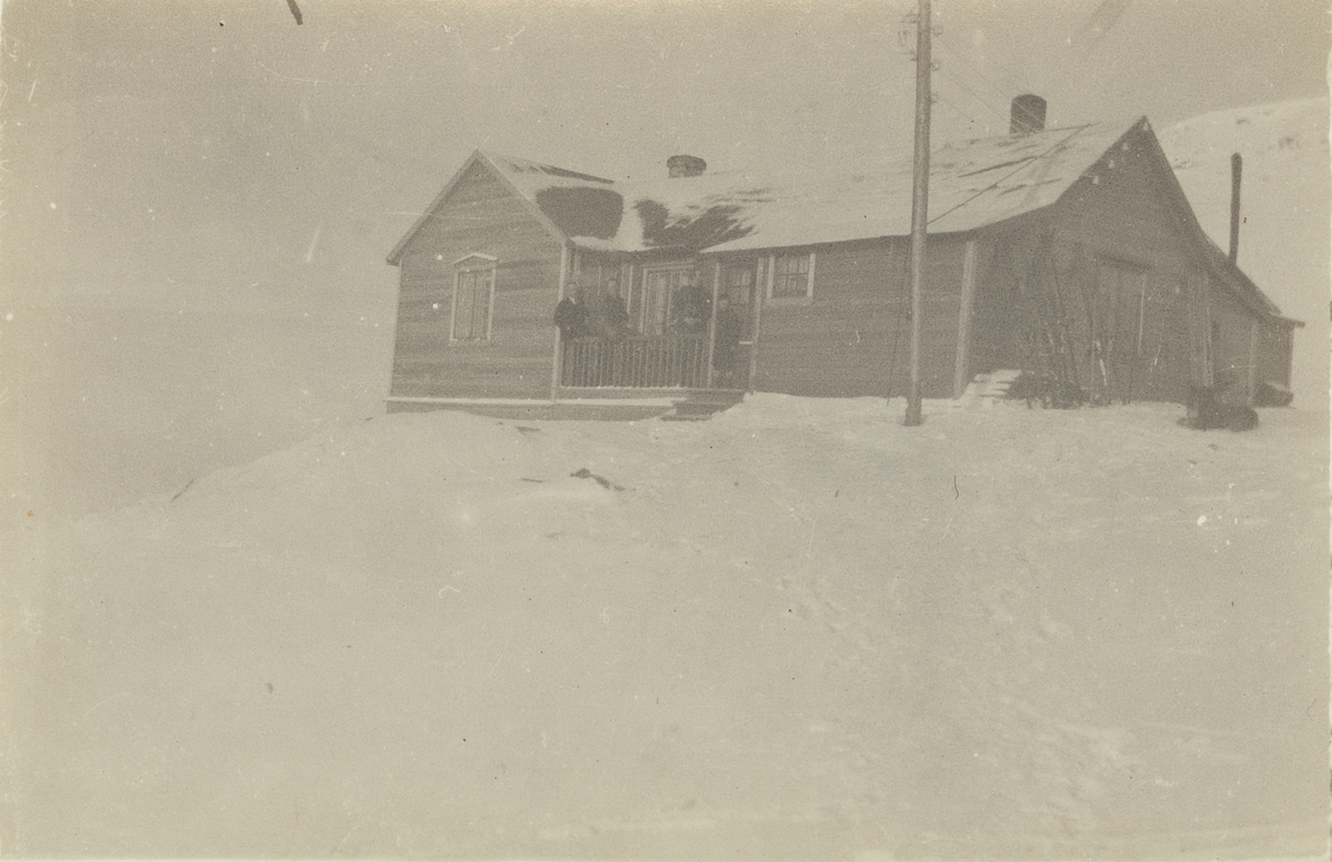 Fotografi från expedition till Spetsbergen. Motiv av några personer som står på verandan till ett trähus som ligger i ett snölandskap. Bilden är troligen tagen vid Sveagruvan.