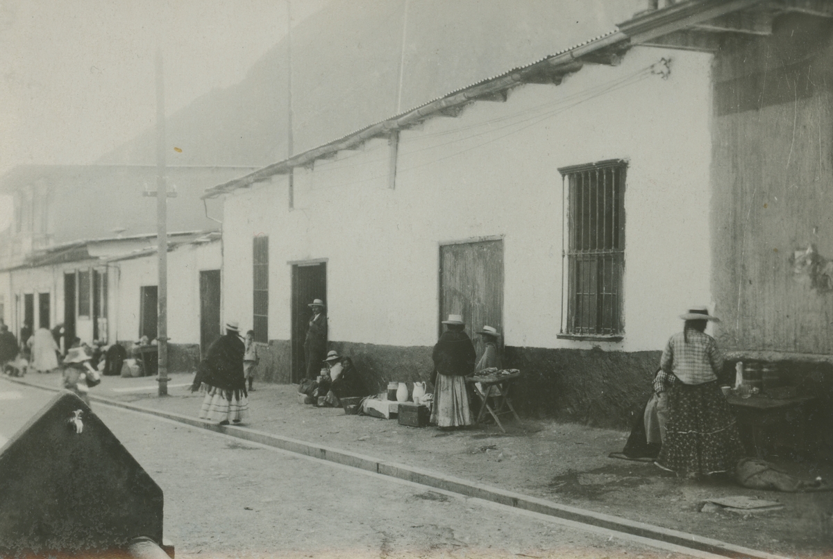 Fotografi från expedition till Peru 1920. Motiv av gata och hus i liten by. På trottoaren syns ett antal kvinnor.