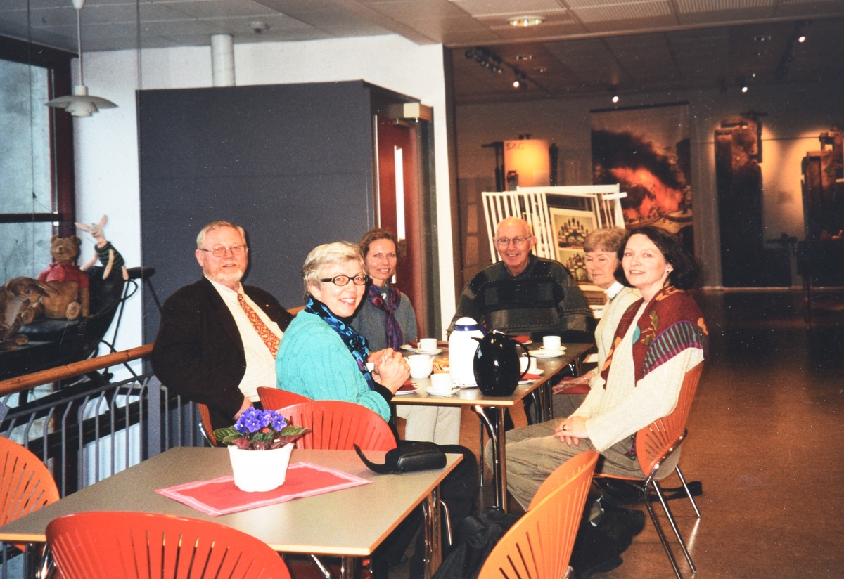 To menn og fire kvinner sit rundt eit bord med te- og kaffikannar og kaffikoppar på bordet. Utstilling skimtas i bakgrunn.