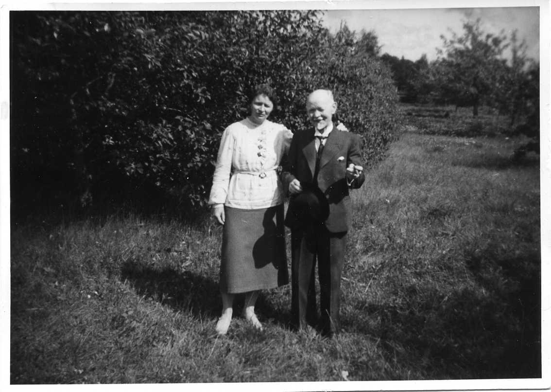Ett äldre par står intill varandra i en trädgård. Mannen har en cigarill e motsv i en hand och en hatt i den andra.