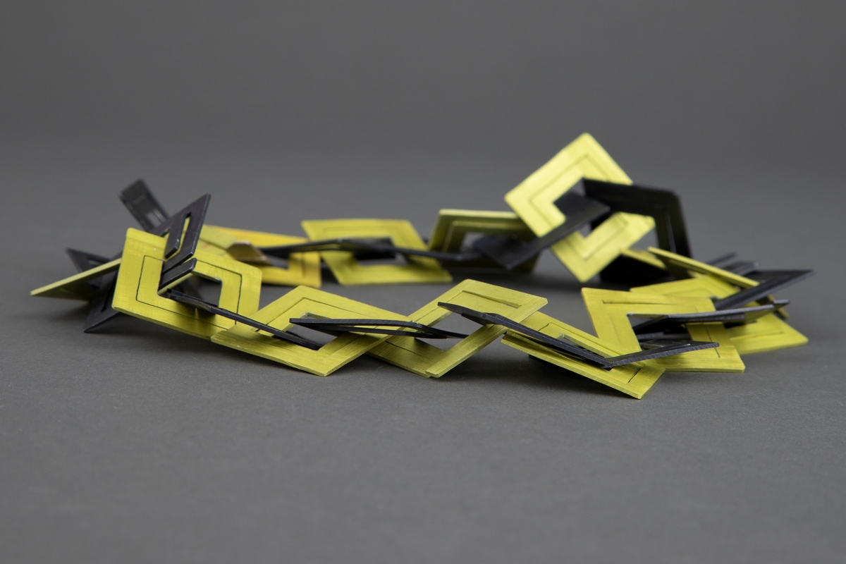 Halskjede av eloksert aluminium, tredd sammen av flate kvadrater i svart og gulgrønt. Hvert kvadrat har en meanderlignende form og er åpen i midten. Halskjedet har et kvadratisk lås i sølv.
