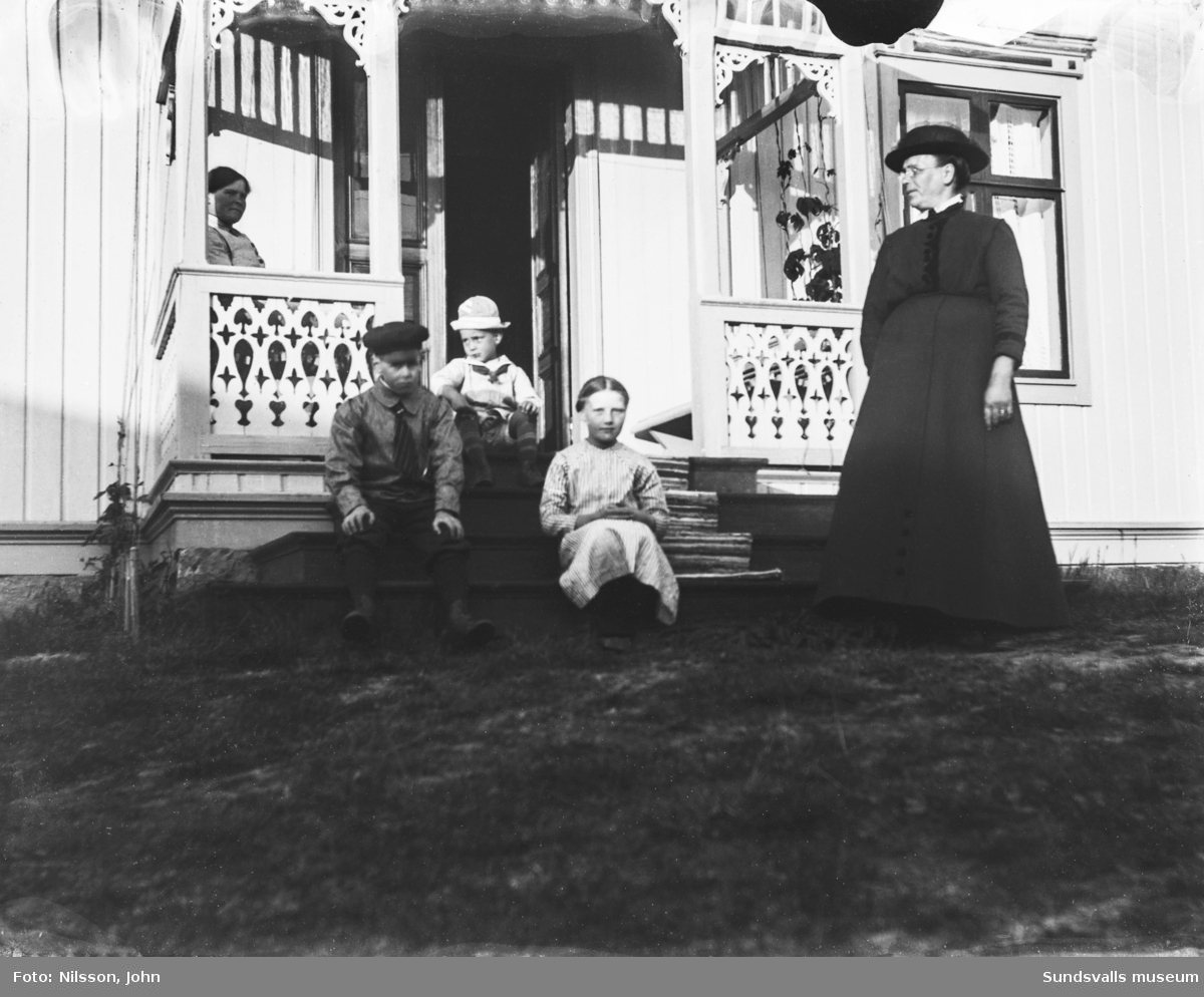 Två bilder med två kvinnor och tre barn på en brokvist med snickarglädje. Huset är ljust målat. Båda bilderna är skadade i överkanten.
