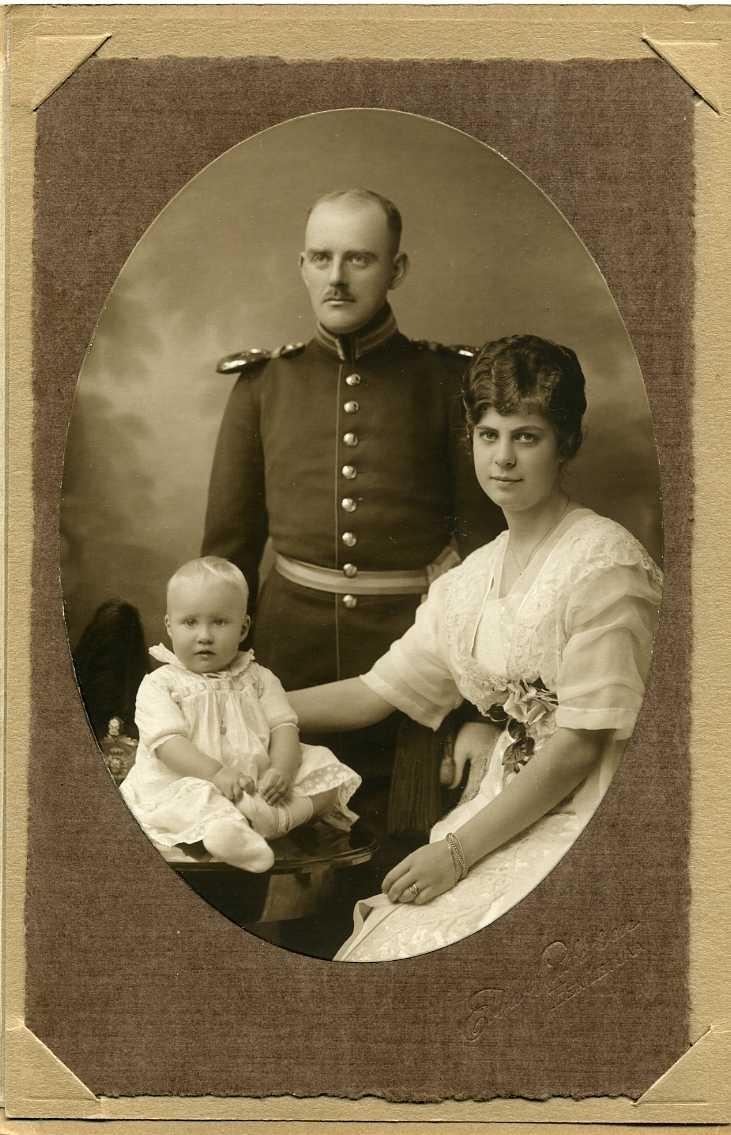 En man i militär uniform står intill en kvinna i ljus klänning med spets. Hon håller höger hand om ryggen på ett spädbarn som sitter på ett bord.
