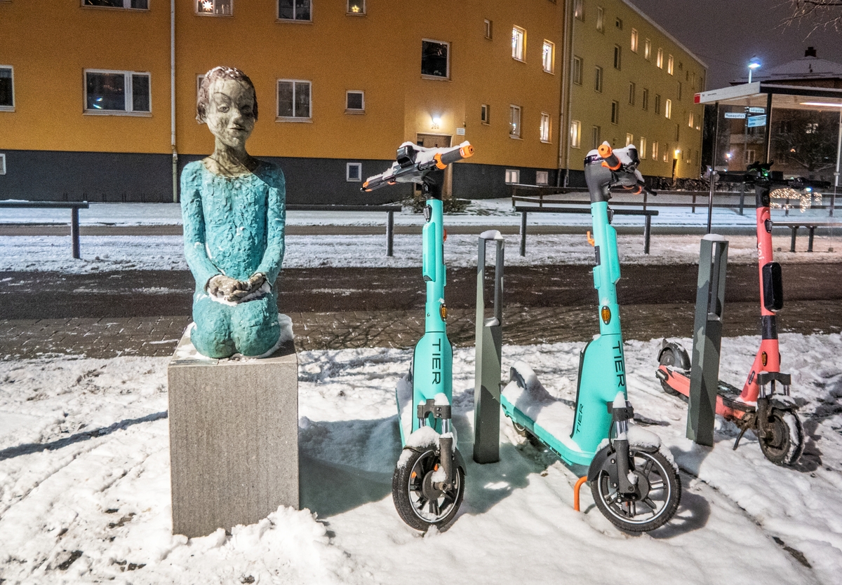 Knäsittande flicka. Skulptur av Lena Cronqvist, 2019. Skulpturen finns på Timmermanstorget i Linköping. Bredvid skulpturen ser man elsparkcyklar parkerade.