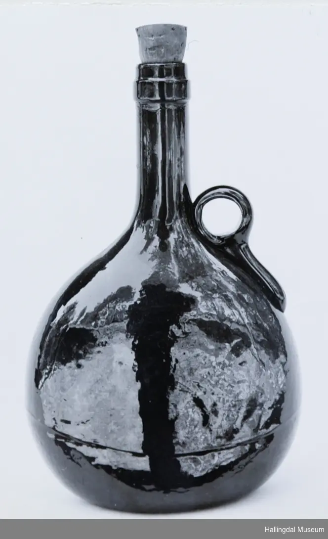 Selve flaske er tilnærmet sirkelrund fra siden, med ovalt tverrsnitt. Tuten sylindrisk. I overgangen til tuten er en rund kork.
6/6-68. Arne L. Christensen.