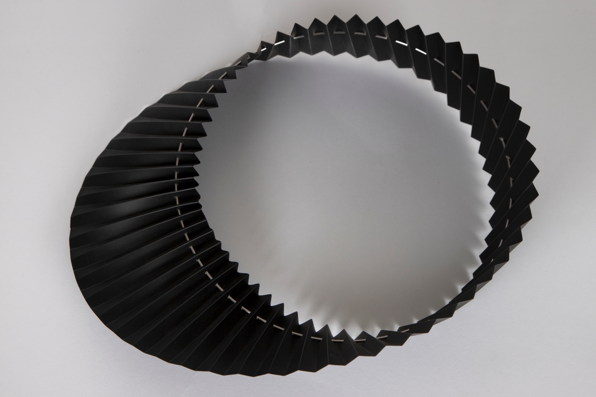 Halskrage av foldelagt svart PVC som er tredd på en sirkelrund stålring og er noe vridd i forhold til denne.