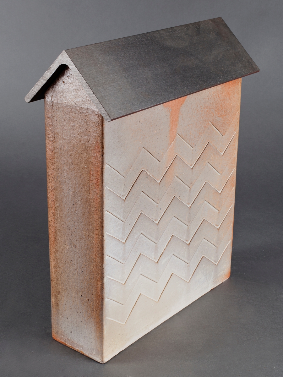 Kiste i hvitglasert steingods med "tak" av stål. Kisten har form som en bygning med smal rektangulær grunnflate og spisst tak. Lavt relieff på langsidene med repetitivt sikksakkmønster. Svakt røde skjolder i overflaten og brun, blank glasur i gavlene.