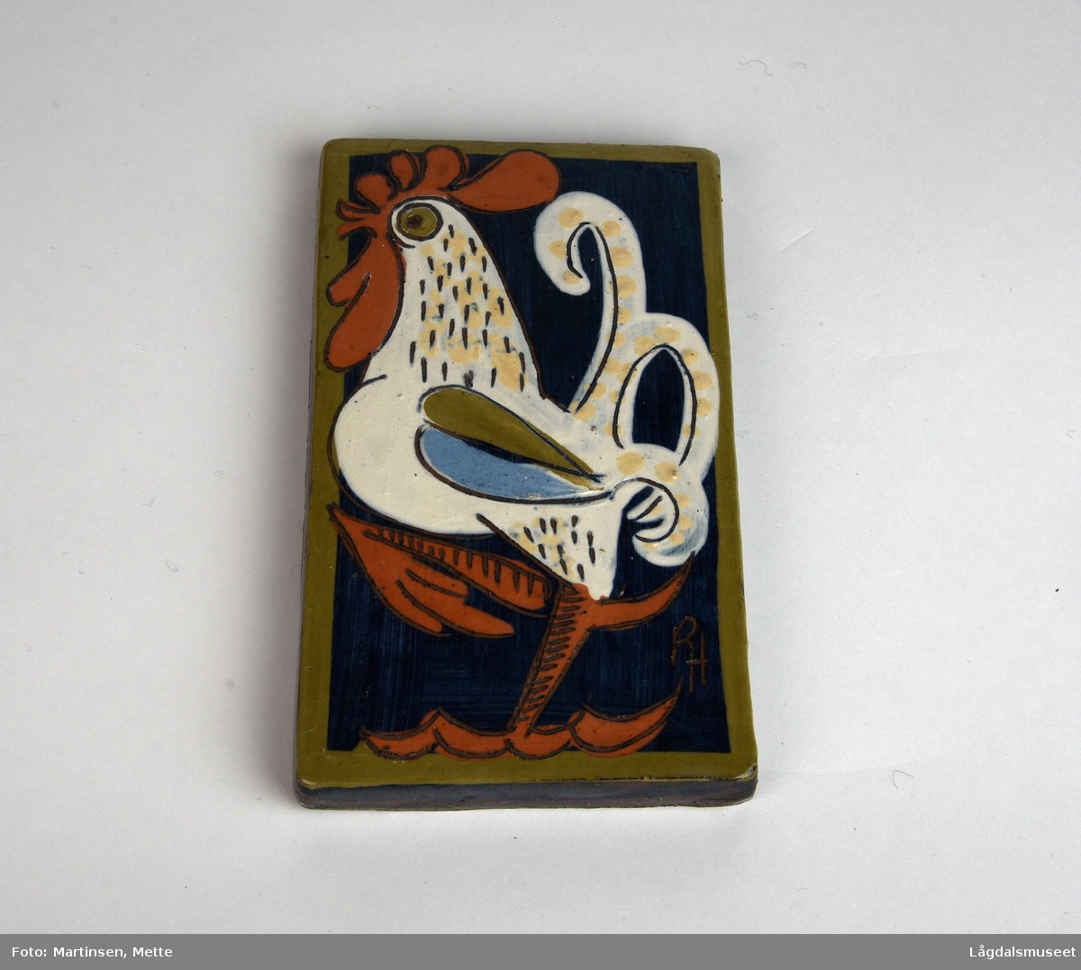 Rektangulær keramikkflis. Motivet er en hvit hane stående på ett bein. Blå bakgrunn og innrammet av en brun kant.