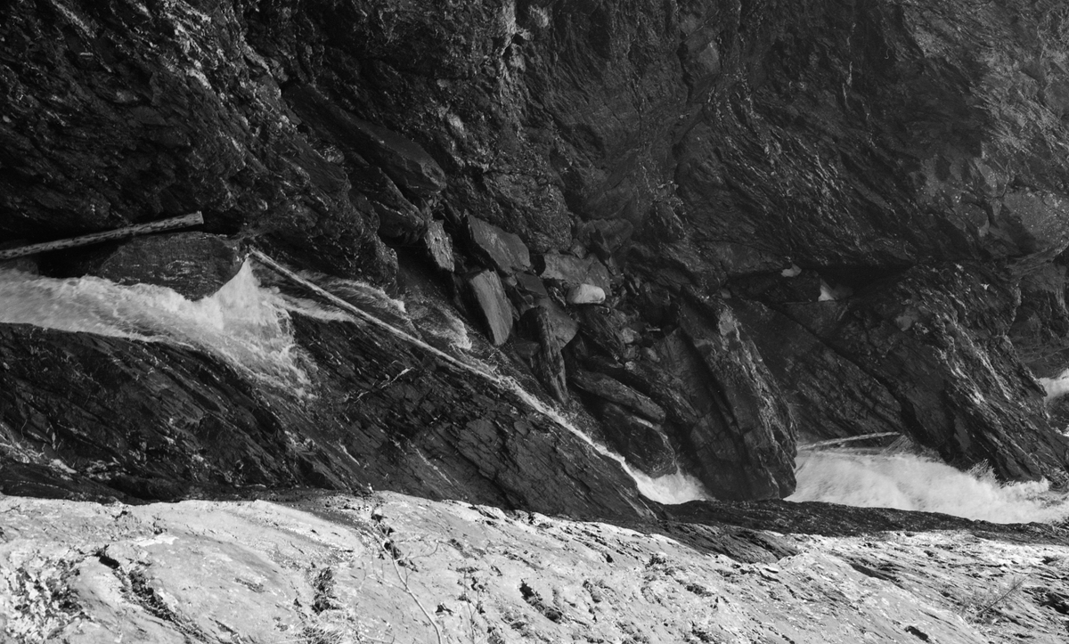 Fra den nedre delen av Kamfossen i Vinstravassdraget i Midt-Gudbrandsdalen, fotografert fra en bergskrent ned mot elveløpet, som renner i ei kløft i fjellet, på dette fotografiet renner fra venstre mot høyre. Kamfossen ligger der Vinstra renner ut av sjøen Olstappen, i Skåbu i Nord-Fron kommune. Fossen har et samlet fall på drøyt 40 meter, riktignok fordelt over flere terskler. Det er åpenbart at det var komplisert å drive tømmerfløting på en elvestrekning som denne. Her var det vanskelig og farefullt for mannskapene å ta seg ned til elveløpet dersom tømmeret satte seg fast. Olstappen ble regulert som magasin for kraftproduksjon i perioden 1953-55. Dette fotografiet er tatt cirka ti år seinere.