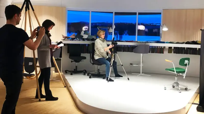 Her der vi tre personer i arbeid i et utstillingsrom. Ei sitter å tar bilde av en stol som er plassert på ei lita scene. Bak henne står ei dame som noterer, og en mann som tar bildet med et mobilkamera.