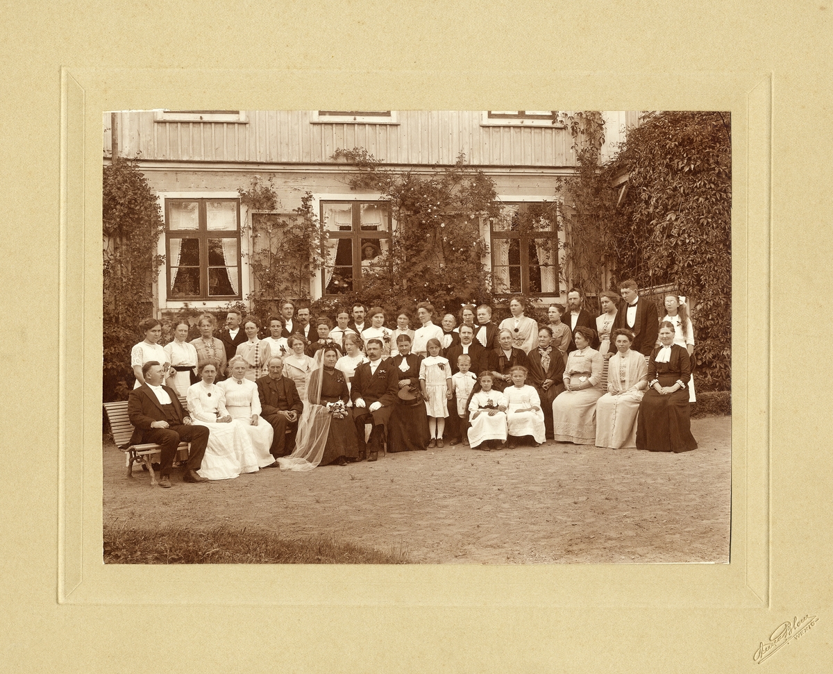 Bröllopsgäster med brudpar och präst, någonstans i Växjö, ca 1912. 
Man har samlats framför bostadshuset i en del av trädgården.
Ulriksberg (?), Östregård (?).