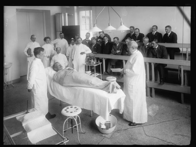 Sort hvitt bilde av en mann som ligger på operasjonsbordet med glede over seg. Mange leger står rundt i hvite frakker.