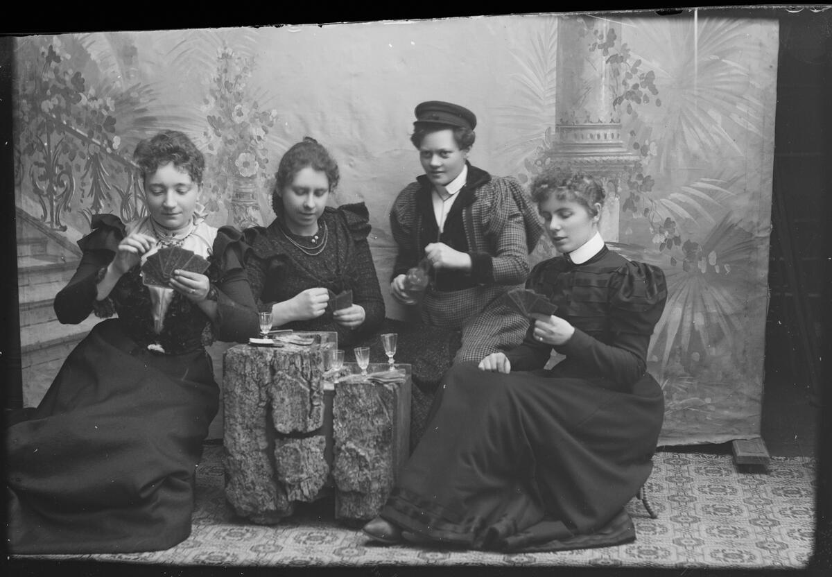Fire kvinner sitter rundt en steinsetting i fotostudio, opptatt med kortspill. De har kjoler på, og stettglass og en liten flaske. Svart-hvitt fotografi.