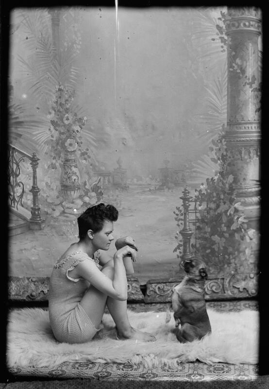 En kvinne med kort hår og ullundertøy sitter på en skinnfell sammen med en hund. Hunden og kvinnen etterligner hverandres positur, med labbene og hendene opp foran seg. Svart-hvitt fotografi.