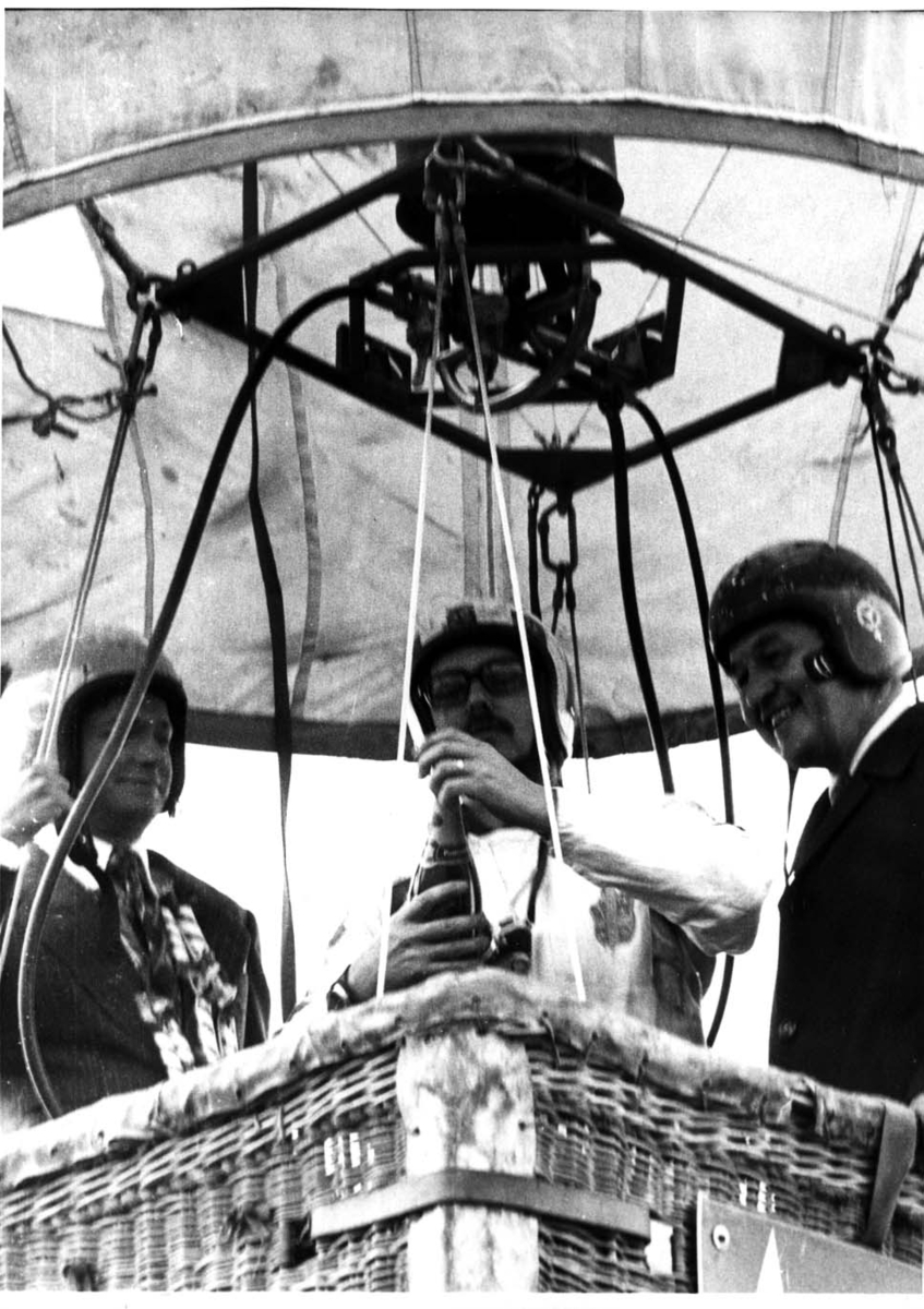 Tre män med hjälm på huvudet står i en ballongkorg. Mannen i mitten öppnar en flaska mousserat av något slag.