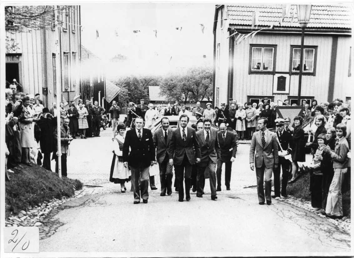 En grupp män på väg uppför en backe på väg till Andréemuseet i Gränna. En kvinna i folkdräkt går bakom dem. En folksamling kantar deras väg