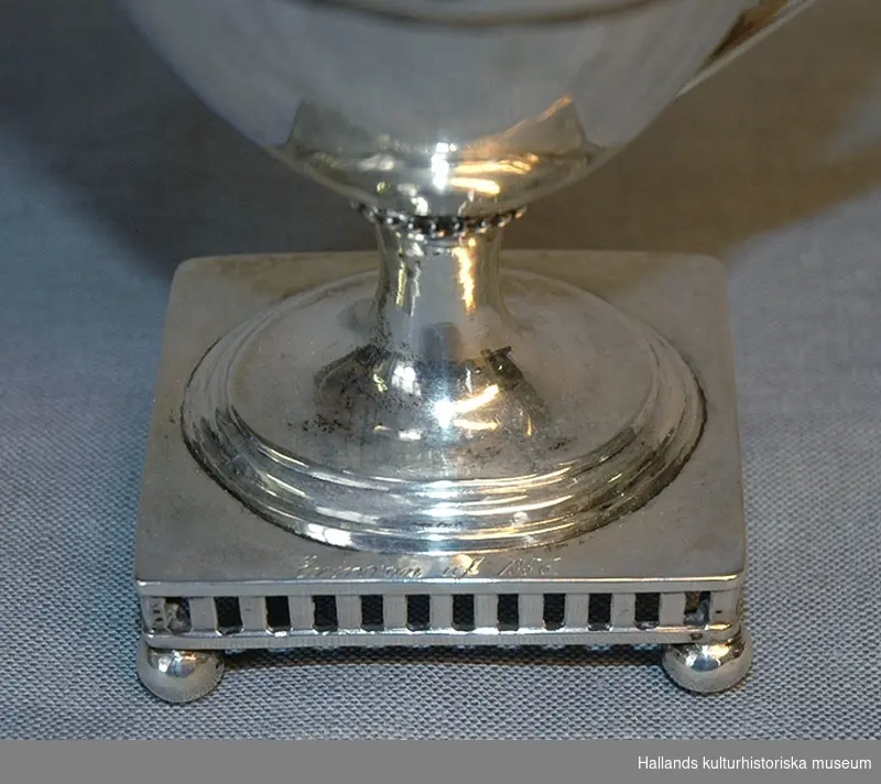 Gustaviansk gräddsnipa av silver. Genombruten fyrkantig fot med kulfötter. Inpunsad vinranksdekor. Stämplar: Stockholms stads stämpel; Tre kronor; "R 2", "GH". På fotplattan ingraverat: "Erinran af 1856."