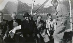 Seks menn ombord i en båt. To røyker pipe. En har en hund ve