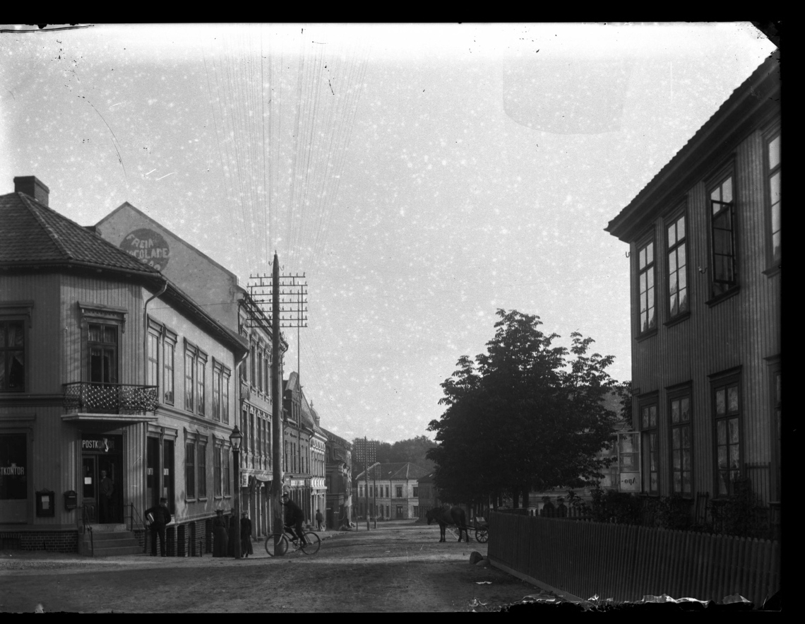 Storgata i Horten, med torvet ned til høyre. Mann på sykkel og gående mennesker ute i gaten. Reklame for Freia sjokolade.