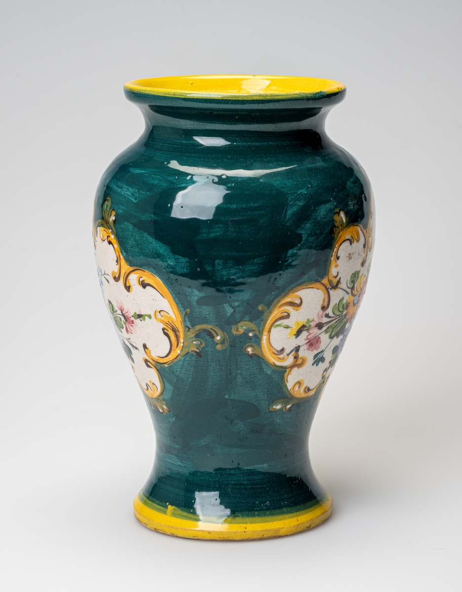 En urneformet vase av keramikk. Kanten langs foten og randen er malt i en klar gulfarge. Ellers er vasen grønnmalt. På hver side av vasen er det et blomstermotiv med blomster i gult, blått, rosa og brunt med grønne blader på hvit bakgrunn. Motivet er omkranset av enkle akantusblader i gult og grønt med detlajer i brunt. Innvendig er vasen hvit/gråhvit. På undersiden er det risset inn tekst (produsent og produksjonsland), se "Påført tekst/merker".